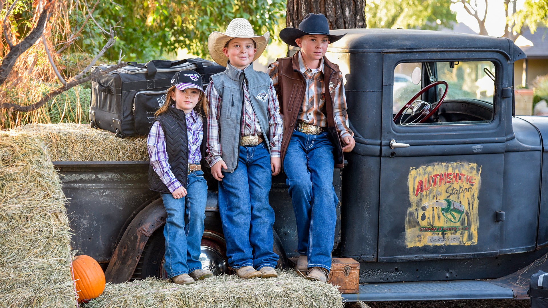 Three little cowboys and cowgirls wearing western wear from Cowboy & Cowgirl Hardware standing infant of an old truck with haystacks