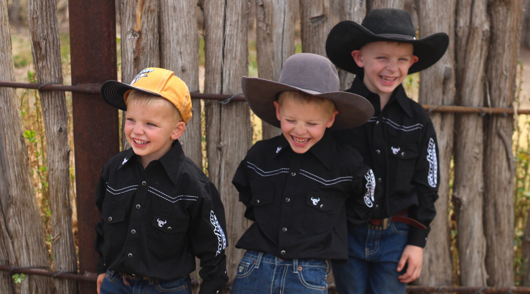 Young cowboys laughing as they lean against a fence wearing matching family sets from Cowboy Hardware.
