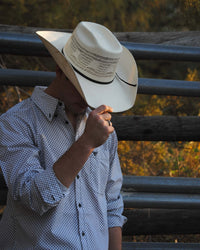 Cowboy holding front of cowboy hat looking down leaning on fence wearing Cowboy Hardware's white puzzle star long sleeve western shirt