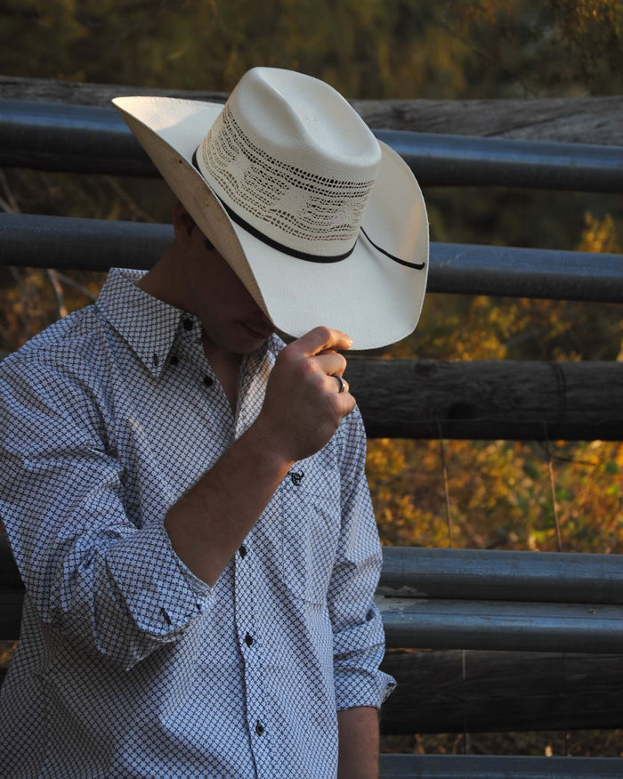 Cowboy holding front of cowboy hat looking down leaning on fence wearing Cowboy Hardware's white puzzle star long sleeve western shirt