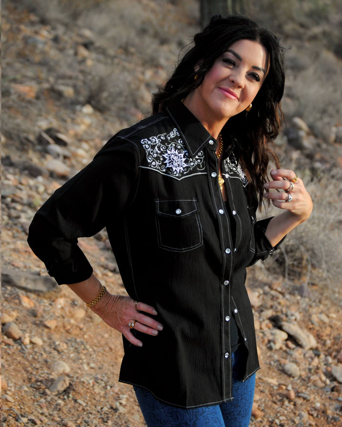 Lady standing in desert smiling smiling at camera wearing Cowgirl Hardware's black vintage embroidered long sleeve solid western shirt