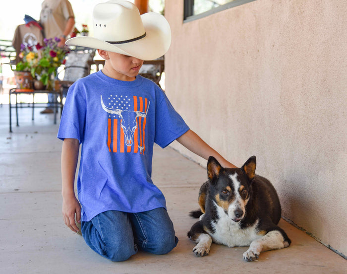 Little cowboy sitting next to his dog petting him on the back patio wearing Cowboy Hardware's youth boys heather blue with orange skull flag short sleeve tee