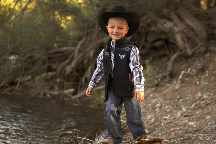 Little cowboy smiling next to creek wearing western outfit including Cowboy Hardware's tonal aztec accent black poly shell vest