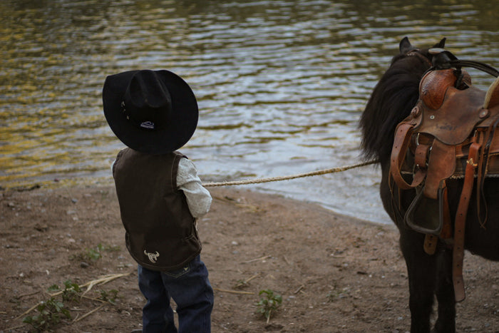 Little cowboy standing next to creek with horse wearing boy's Cowboy Hardware dark chocolate live free tech woodsman vest