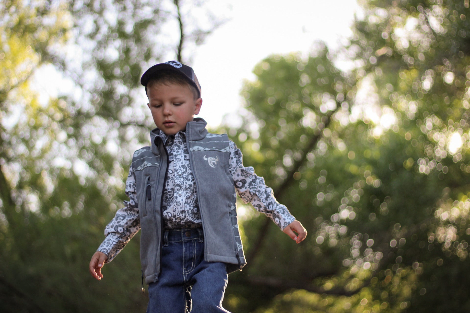 Little cowboy wearing boy's Cowboy Hardware tonal aztec accent grey poly shell vest walking through the woods