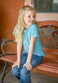 Little cowgirl sitting on front porch bench wearing Cowgirl Hardware's infant and toddler girls turquoise acid wash crew neck shorts sleeve top