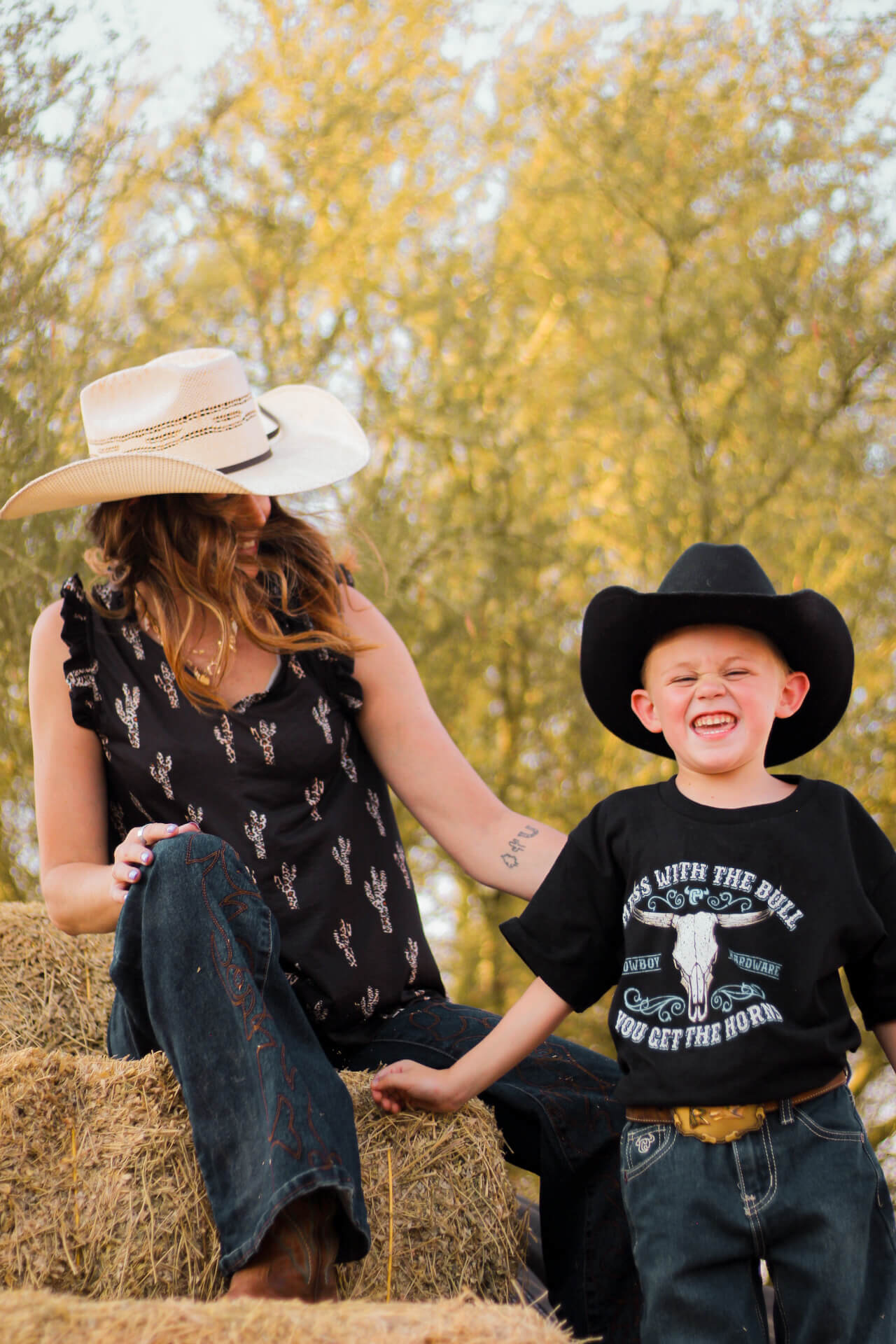 Woman wearing Cowgirl Hardware Leopard Saguaro Black Ruffle V-neck Tank with a little cowboy hanging out on a pile of hay.