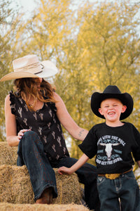 Woman wearing Cowgirl Hardware Leopard Saguaro Black Ruffle V-neck Tank with a little cowboy hanging out on a pile of hay.